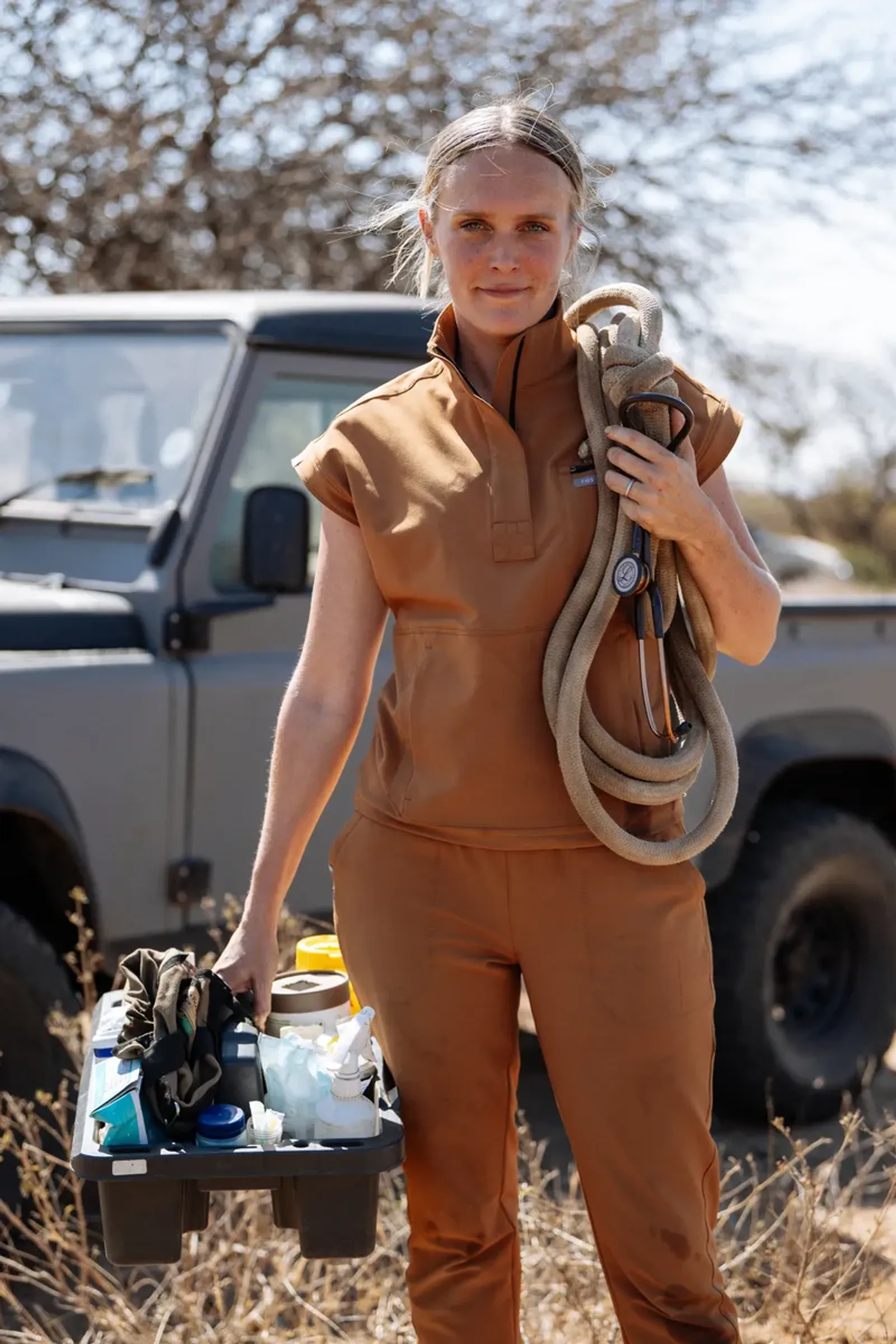 Chloe in vet scrubs by a truck, a rope and stethoscope over her shoulder, carrying a tray of supplies